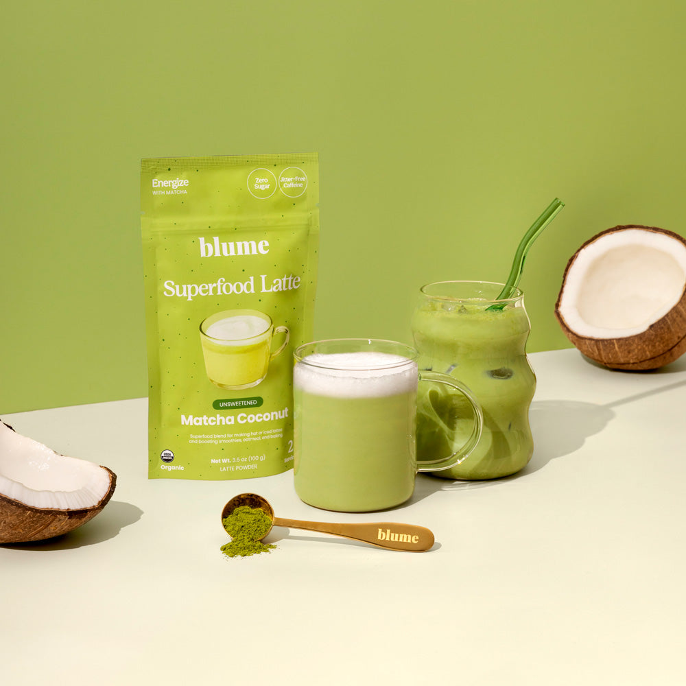 A pouch of Blumes Matcha Coconut Blend, antioxidant-rich, is showcased beside a glass mug latte and iced latte jar. A spoon with matcha powder and two coconut halves complement the scene on a calming green backdrop.